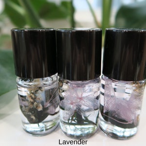 May include: Three small glass bottles with black lids, each containing a different type of dried flower preserved in clear liquid. The flowers are lavender, white, and a light purple.