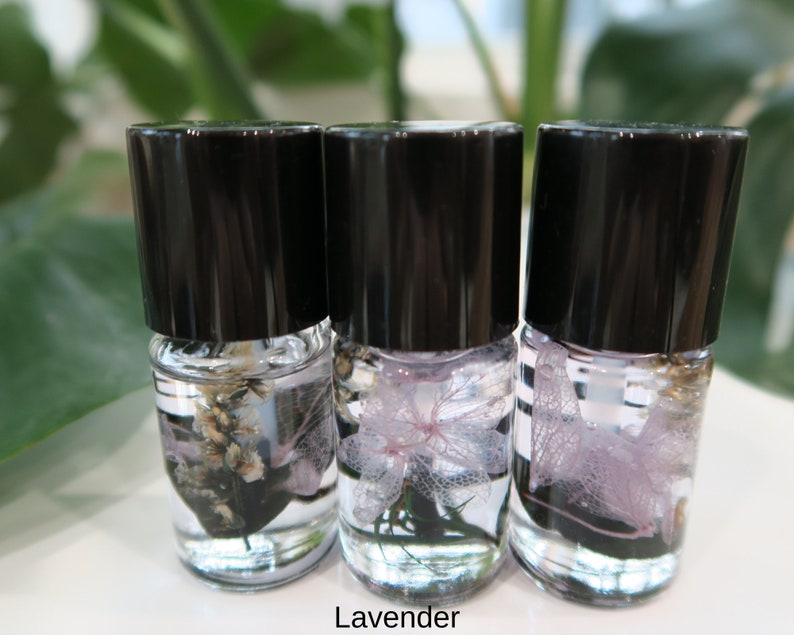 May include: Three small glass bottles with black lids, each containing a different type of dried flower preserved in clear liquid. The flowers are lavender, white, and a light purple.