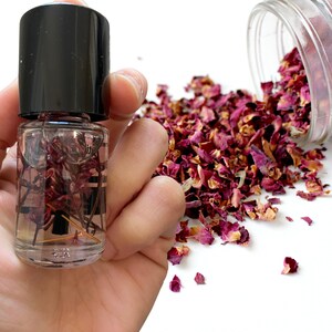 May include: A small glass bottle filled with dried rose petals and a clear liquid. The bottle has a black cap and is held in a hand. Dried rose petals are scattered on a white surface.