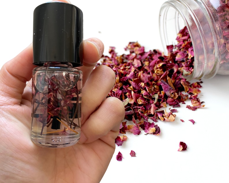 May include: A small glass bottle filled with dried rose petals and a clear liquid. The bottle has a black cap and is held in a hand. Dried rose petals are scattered on a white surface.