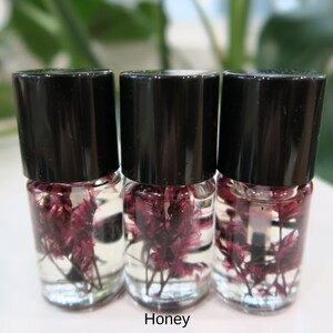 May include: Three small glass bottles with black lids filled with clear liquid and dried purple flowers. The bottles are arranged in a row on a white surface. The word "Honey" is printed below the bottles.