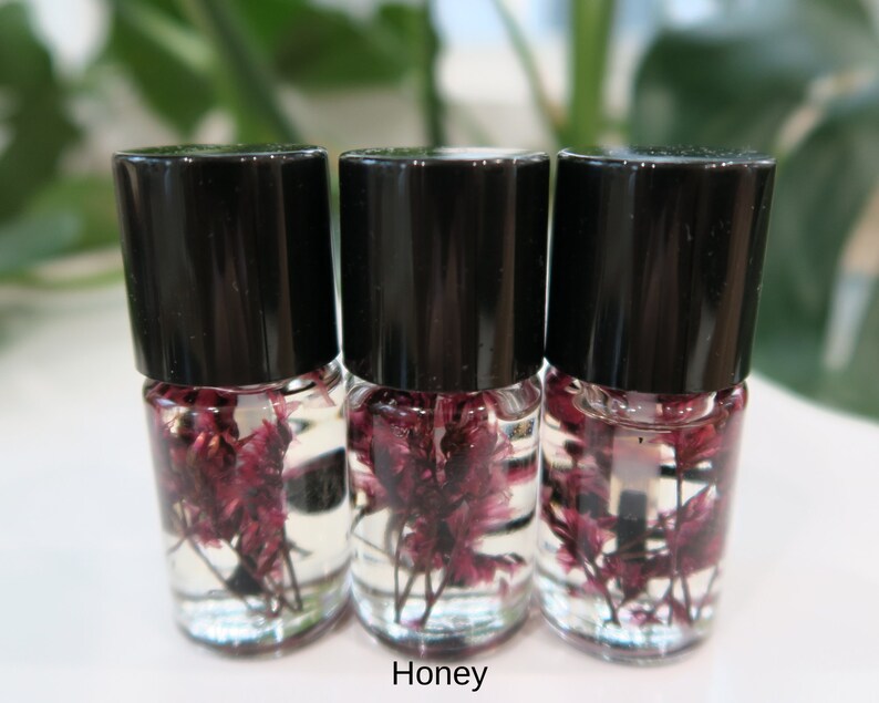 May include: Three small glass bottles with black lids filled with clear liquid and dried purple flowers. The bottles are arranged in a row on a white surface. The word "Honey" is printed below the bottles.
