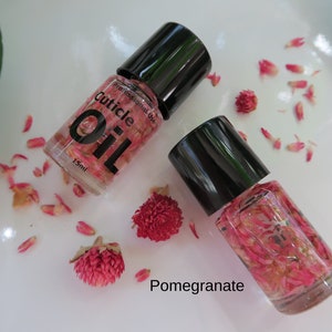 May include: Two small bottles of cuticle oil with dried flowers inside. The bottles are clear glass with black caps. The label on one bottle reads "Professional Use Cuticle Oil 15ml". The text "Pomegranate" is written below the bottles.
