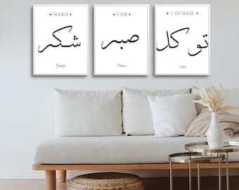 May include: Three white framed prints with black calligraphy in Arabic script. The prints are arranged horizontally on a white wall. The text in the prints reads "Shukr", "Sabr", and "Tawakkul" in Arabic. The English translations of the words are "Grateful", "Patience", and "Trust" respectively.