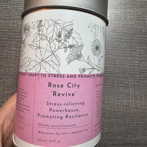 Op de afbeelding: Een cilindrische metalen container met een zilveren deksel en een wit etiket. Het etiket bevat botanische illustraties en de tekst "Rose City 'Revive' Stress-relieving Powerhouse, Promoting Resilience". De container bevat 355 ml.
