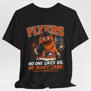 Può includere: T-shirt nero con la mascotte dei Philadelphia Flyers, Gritty, che indica lo spettatore. La maglietta presenta il logo dei Flyers e la scritta "NO ONE LIKES US. WE DON'T CARE." in arancione e bianco. È incluso anche il testo "BROAD STREET BULLIES SINCE '67".