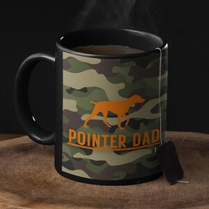 May include: A black coffee mug with a camouflage pattern and the text "POINTER DAD" in orange. The mug has a black handle and is filled with coffee. There is steam rising from the coffee.