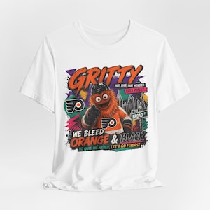 Op de afbeelding: Wit T-shirt met een cartoon illustratie van Gritty, de mascotte van de Philadelphia Flyers. Het ontwerp bevat de tekst "GRITTY" en "WE BLEED ORANGE & BLACK". Het logo van de Flyers is ook aanwezig.