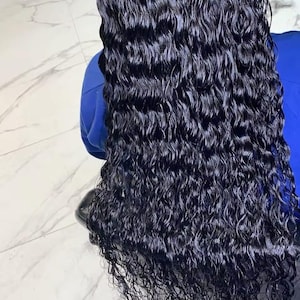Könnte beinhalten: Lange, dunkle, wellige Haare mit einem nassen, glänzenden Aussehen. Das Haar fällt herab und zeigt seine natürlichen Locken und Textur. Das Haar hat eine tiefschwarze Farbe, die das Licht reflektiert und einen glänzenden Effekt erzeugt. Es scheint sich um eine Perücke oder Haarverlängerung zu handeln.
