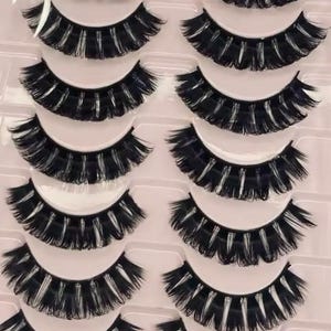 Könnte beinhalten: Ein Set mit 10 Paaren schwarzer künstlicher Wimpern in einem durchsichtigen Plastikbehälter. Die Wimpern sind in Reihen angeordnet und bereit zum Anbringen.