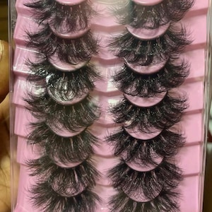 Op de afbeelding: Een set van zes paar valse wimpers in een doorzichtige plastic doos met roze scheidingswanden. De wimpers zijn zwart en hebben een luchtige, gelaagde look.