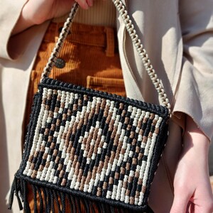 May include: A macrame crossbody bag with a geometric pattern in black, brown, and white. The bag has a black fringe along the bottom and a woven strap. The bag is held by a person.