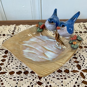 May include: A square ceramic trinket dish with a textured tan surface and a white glazed center. Two blue birds with brown details are perched on the edge, accompanied by small green leaves and clusters of red berries attached to the rim.