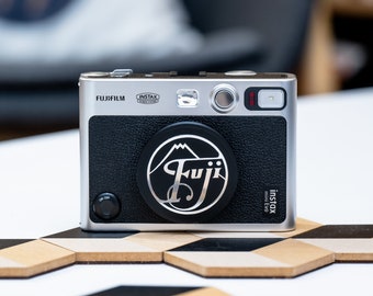 May include: A silver and black Fujifilm Instax Mini Evo instant camera with a black leather-like finish. The camera has a round lens cover with a silver "Fuji" logo and a mountain graphic. The camera also has a red light on the top right corner.