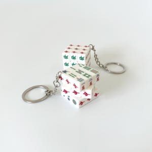 Puede incluir: Dos llaveros en forma de cubo blanco con símbolos de fichas de Mahjong de colores en cada cara. Cada llavero está unido a una anilla de metal plateado mediante una cadena corta. Los cubos muestran varios caracteres y patrones en rojo, verde y azul.