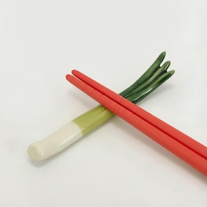 Puede incluir: Un par de palillos chinos rojos descansan sobre un reposapalillos de cerámica con forma de puerro. El reposapalillos es blanco, verde y amarillo. Los palillos chinos son de color rojo brillante. El reposapalillos de puerro es un artículo decorativo.