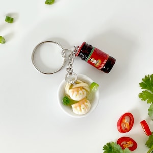 May include: A novelty keyring featuring a miniature bowl of dumplings with noodles, a small red bottle, and a silver keyring. The dumplings are white with a light brown sauce. The bottle is red with a label.