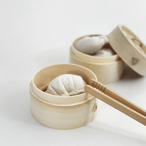 Può includere: Primo piano di due cestini per la cottura a vapore in bambù, ciascuno contenente un raviolo. Un cestino è aperto, rivelando diversi ravioli. Un paio di pinze di bambù sono appoggiate di lato. Lo sfondo è bianco.