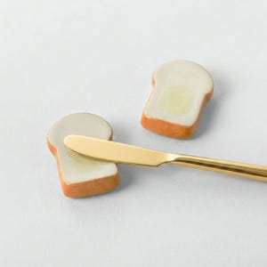 Puede incluir: Un cuchillo de mantequilla dorado descansa sobre dos mantequilleras con forma de pan. Las rebanadas de pan tienen un centro blanco y una corteza dorada, con un cuadrado de mantequilla amarilla en el centro. El fondo es blanco.