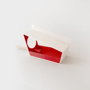 Puede incluir: Un comedero para pájaros moderno con exterior blanco e interior rojo vibrante. El comedero presenta un diseño rectangular con un techo inclinado y una abertura circular. Una percha de madera se extiende desde el lateral.