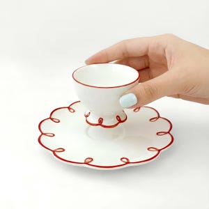 May include: A white teacup and saucer set with red trim. The teacup has a rounded bowl and a pedestal base. The saucer has a scalloped edge with red detailing. The set is designed for serving tea or coffee.