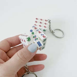 Könnte beinhalten: Ein weißer Mahjong-Würfel-Schlüsselanhänger mit roten, grünen und blauen Markierungen. Der Schlüsselanhänger hat einen silbernen Metallring und eine Kette. Der Würfel wird in einer Hand mit hellblauem Nagellack gehalten.