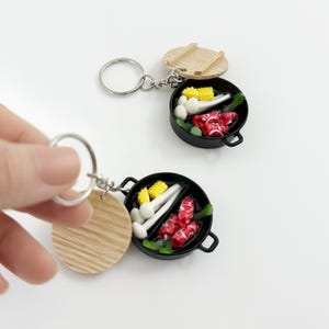 Puede incluir: Dos llaveros de comida en miniatura. Cada llavero tiene una sartén negra con tapa, llena de maíz, champiñones y carne roja. Los llaveros tienen un anillo plateado y un disco de madera.