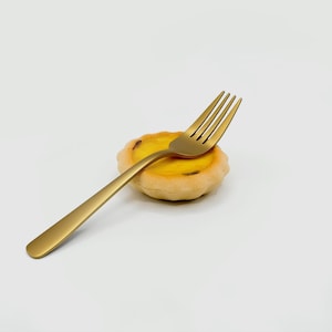 Può includere: Una forchetta color oro è appoggiata su una piccola pasta rotonda con un ripieno di crema pasticcera gialla. La forchetta ha un manico lungo e sottile e quattro rebbi. La pasta ha una crosta dorata.