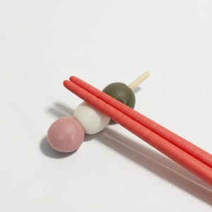Puede incluir: Primer plano de dango japonés, una bola de masa dulce, con palillos rojos. Los dangos son rosas, blancos y verdes, ensartados en un palito de madera. Los palillos son de color rojo vibrante.