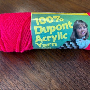 Puede incluir: Una madeja de hilo acrílico DuPont 100% de color rosa brillante. La etiqueta presenta a una mujer con cabello castaño largo y una camisa blanca con un diseño estampado en blanco y negro. La etiqueta también dice "100% DuPont Acrylic Yarn, 4 ply Knitting Worsted."