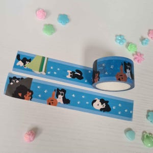 May include: Blue washi tape with a repeating pattern of black cats wearing glasses and brown dogs. The tape is rolled up on a blue spool.