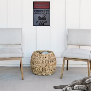 May include: A warning sign that reads "WARNING PROPERTY GUARDED BY ATTACK CAT" is hanging on a white wall. Two light gray chairs with wooden legs are positioned on either side of a woven wicker basket. A gray cat is lying on the floor in front of the chairs.