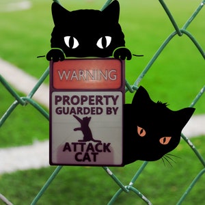 May include: A black cat peeks through a chain link fence with a sign that reads "WARNING PROPERTY GUARDED BY ATTACK CAT".