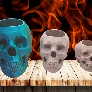 May include: Three skull-shaped planters, one blue, two gray, on a wooden surface. The planters are made of a textured material and have a hollow interior.