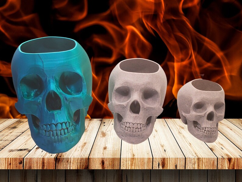 May include: Three skull-shaped planters, one blue, two gray, on a wooden surface. The planters are made of a textured material and have a hollow interior.