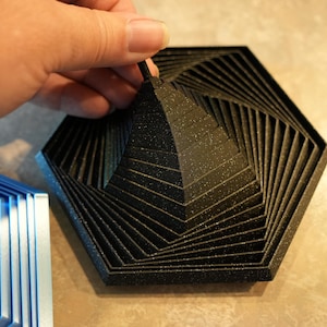 May include: A black, hexagonal, 3D-printed art piece with a pyramid-like structure in the center. The design features layered, angled lines that create a spiral effect. The piece is held by a hand.
