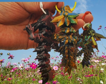 May include: Three origami dragon keychains in different colors: black, gold, and green. The dragons are made of folded paper and have a shiny, iridescent finish. The keychains are hanging from a hand against a background of pink and white flowers.
