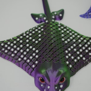 May include: A purple and green stingray with white spots, made of a textured material. The stingray has a 3D design and is a decorative object.