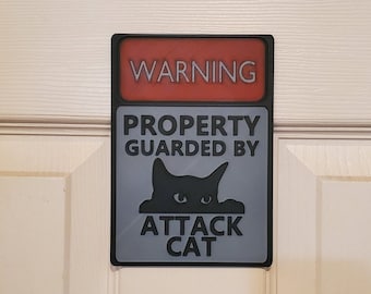 May include: A black and grey sign with the text "WARNING PROPERTY GUARDED BY ATTACK CAT" in white. The sign has a red background with the word "WARNING" in white.