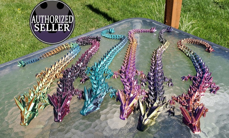 May include: A row of eight iridescent origami dragons in various colors, including green, purple, blue, and gold. The dragons are arranged in a line on a table with a green grassy background.