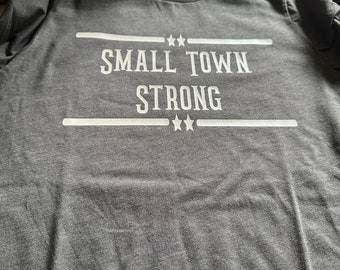 May include: A gray t-shirt with the text "SMALL TOWN STRONG" printed in white on the front. The text is surrounded by two horizontal lines and three stars.