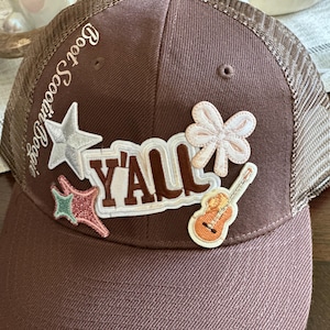 May include: Brown trucker hat with a mesh back. The hat has several patches on the front, including a star, a guitar, a bow, and the word "Y'all". The hat also has the text "Boot Scootin' Boogie" on the side.