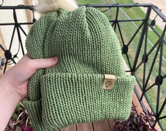 May include: A green knitted beanie with a white pom pom on top. The beanie has a brown leather tag with the text "Craftshop Co."