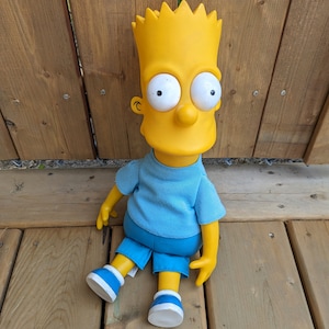 May include: A yellow cartoon character doll with a blue shirt and yellow shorts. The doll has a yellow head with black eyes and a yellow smile.