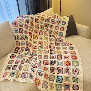 May include: A handmade crochet blanket featuring a repeating granny square pattern. Each square is composed of a central colourful floral motif surrounded by a cream-coloured border, creating a vibrant, multi-coloured textile with a textured finish.