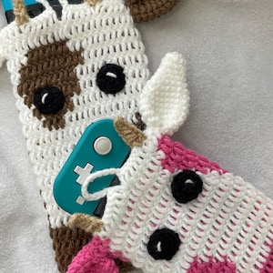 以下が含まれることがあります： ゲームデバイス用の2つの手編みケース。それぞれ牛のデザイン。1つは茶色の斑点のある白、もう1つはピンクのストライプの白。どちらも黒い目と小さな角があります。ターコイズ色のゲームデバイスが部分的に見えます。