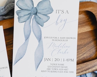 May include: Two baby shower invitations with a watercolor blue bow design and the text "It's a Boy." The invitations are on a wooden tray with a pair of scissors and a blue fabric. The invitations include the baby shower details.