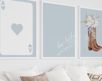 May include: Three framed prints on a white wall above a white couch with brown throw pillows and a brown blanket. The prints are on a light blue background. The left print shows an ace of hearts playing card. The middle print has the text "her lucky charm". The right print shows a brown cowboy boot with white flowers and a blue ribbon.