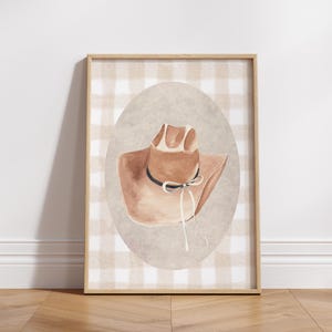 May include: A framed print of a brown cowboy hat with a white ribbon on a gingham background.