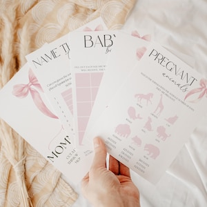 May include: Four pink and white baby shower game cards with pink bows. The cards are titled "Name That Tune", "Baby Bingo", "Mom's Cutest Thinking Spot", and "Pregnant Animals".